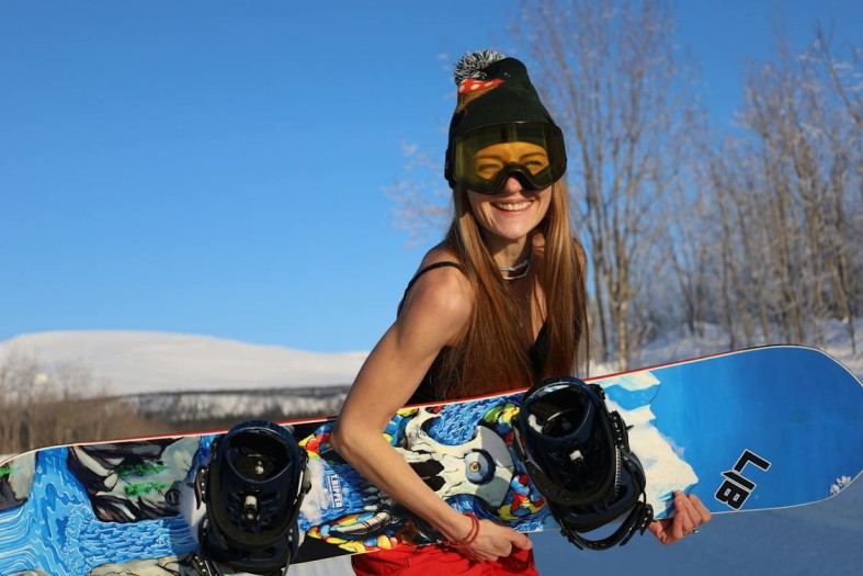 Young Woman in Beanie Hat and Goggles Carrying a Snowboard 