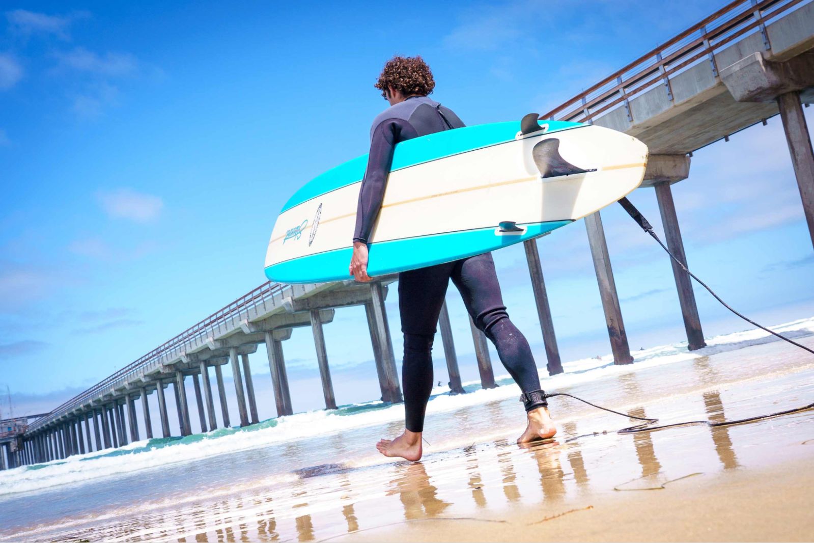 Surfer with a blue surfboard walking along the beach by a pier